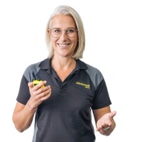 Frau mit kinnlangem, blonden Haar und Brille lächelt in die Kamera. Sie trägt ein Dienst-Shirt der ÖAMTC Fahrtechnik. In ihrer rechten Hand hält sie ein Funkgerat.