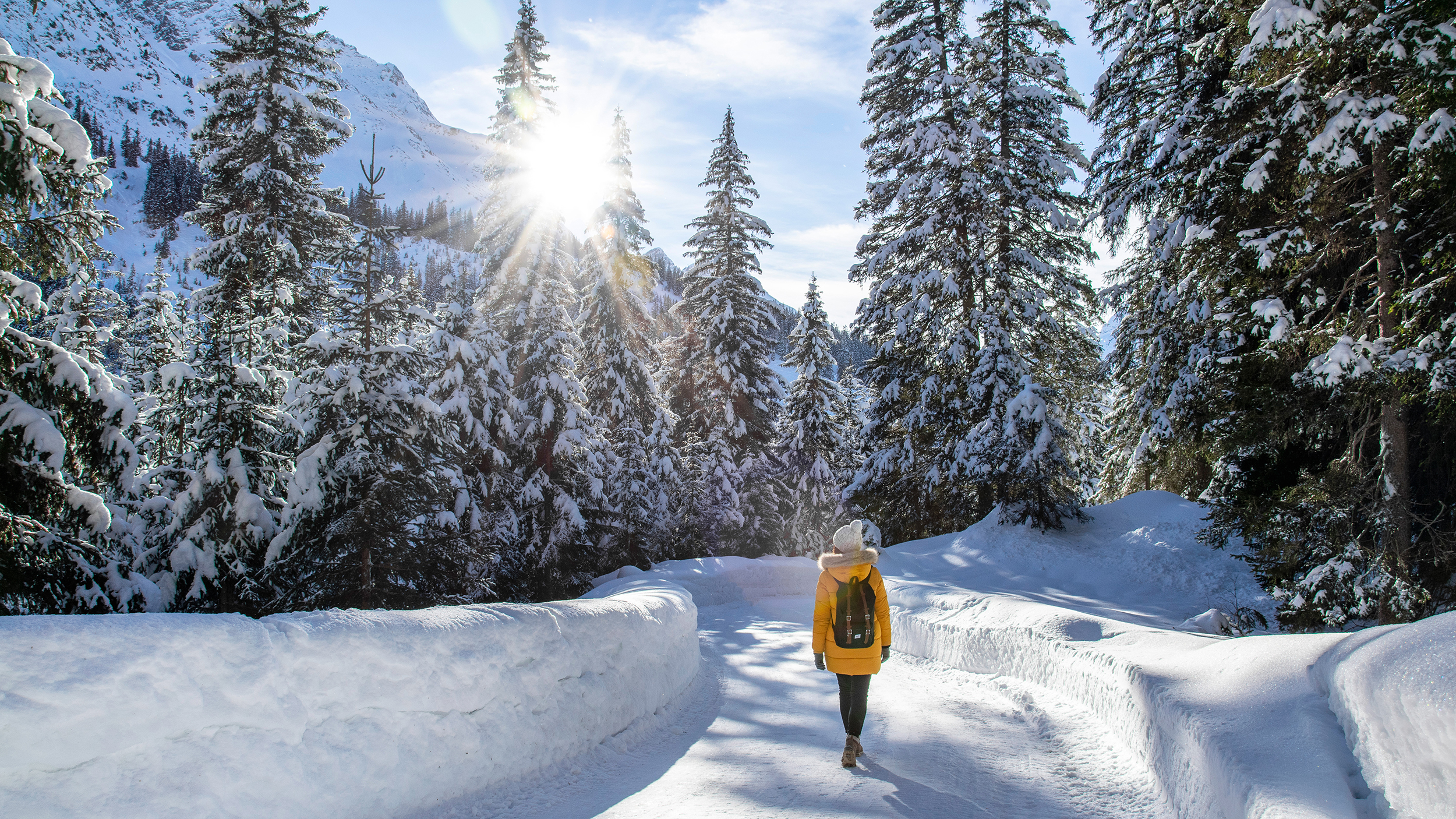 Eine Frau in einem gelben Anorak spaziert durch einen verschneiten Wald. 
