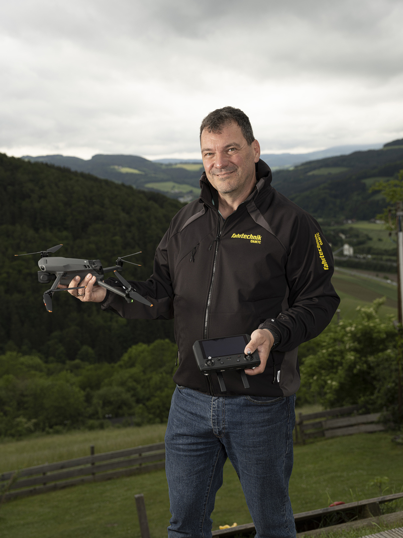 Auf dem Bild sieht man einen Mann vor einer grünen Landschaft. In der Hand hält er eine Drohne. er trägt eine blaue Jean und eine schwarze Jacke, auf der ÖAMTC Fahrtechnik steht.