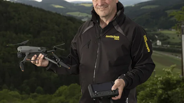 Auf dem Bild sieht man einen Mann vor einer grünen Landschaft. In der Hand hält er eine Drohne. er trägt eine blaue Jean und eine schwarze Jacke, auf der ÖAMTC Fahrtechnik steht.