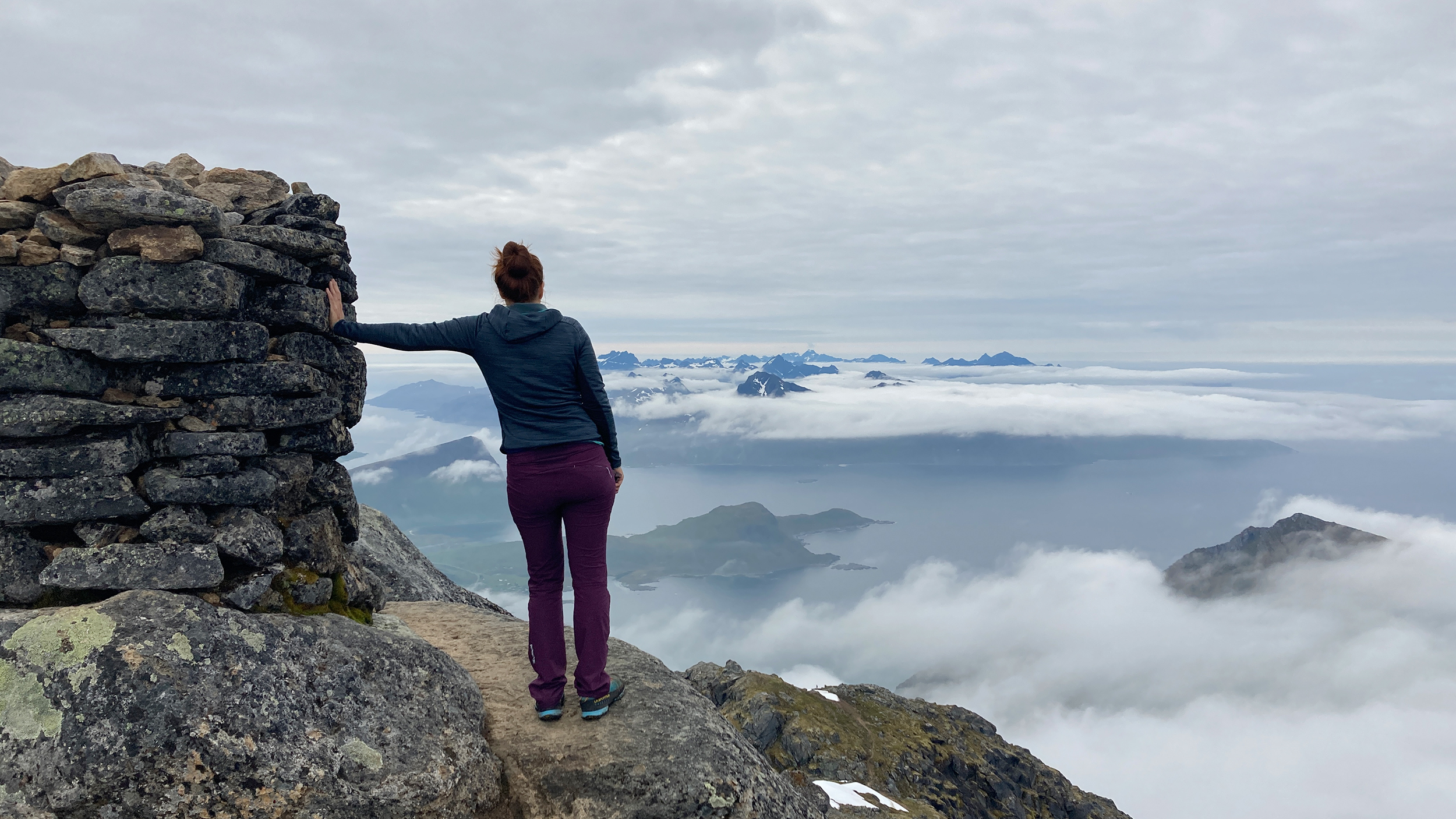 Rückansicht einer Frau, die am Gipfel steht und auf ein wolkenverhangenes Panorama blickt.