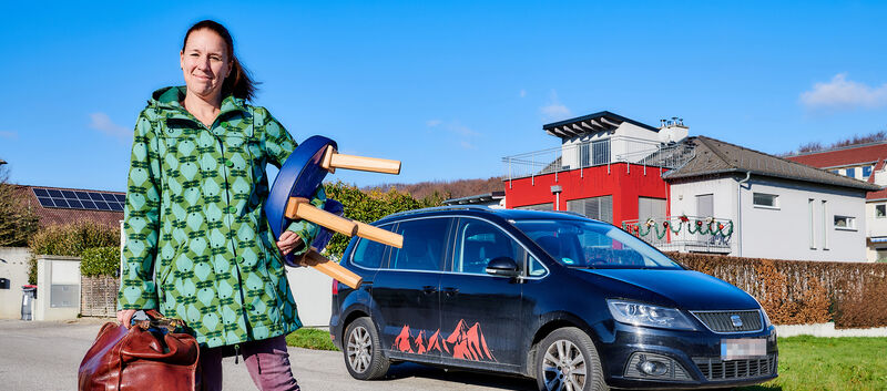 Hebamme Martina Klasz hält ihr Arbeitsgerät: Gebärstuhl und Tasche in der Hand und steht vor ihrem Auto.