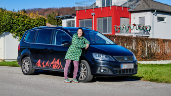 Eine Frau, die Hebamme Martina Klasz, steht an ihr schwarzes Auto gelehnt und lächelt. Im Hintergrund ein Wohnhaus.