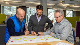 Drei Personen – es handelt sich um einen Verlagsleiter, Chefredakteur und Chef vom Dienst – stehen vor verschiedenen Seitenspiegeln und besprechen sich.