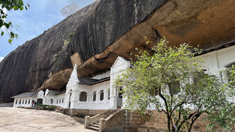 Höhlentempel in Dambulla in Sri Lanka. Aufnahme von außen, man sieht ein weißes Gebäude, dahinter Felsen.