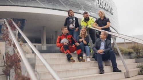 Sechs Mitarbeiter und MItarbeiterinnen in unterschiedlichen Dienstkleidungs-varianten sitzen und stehen abwechselnd auf der Treppe vor dem Mobilitätszentrum.