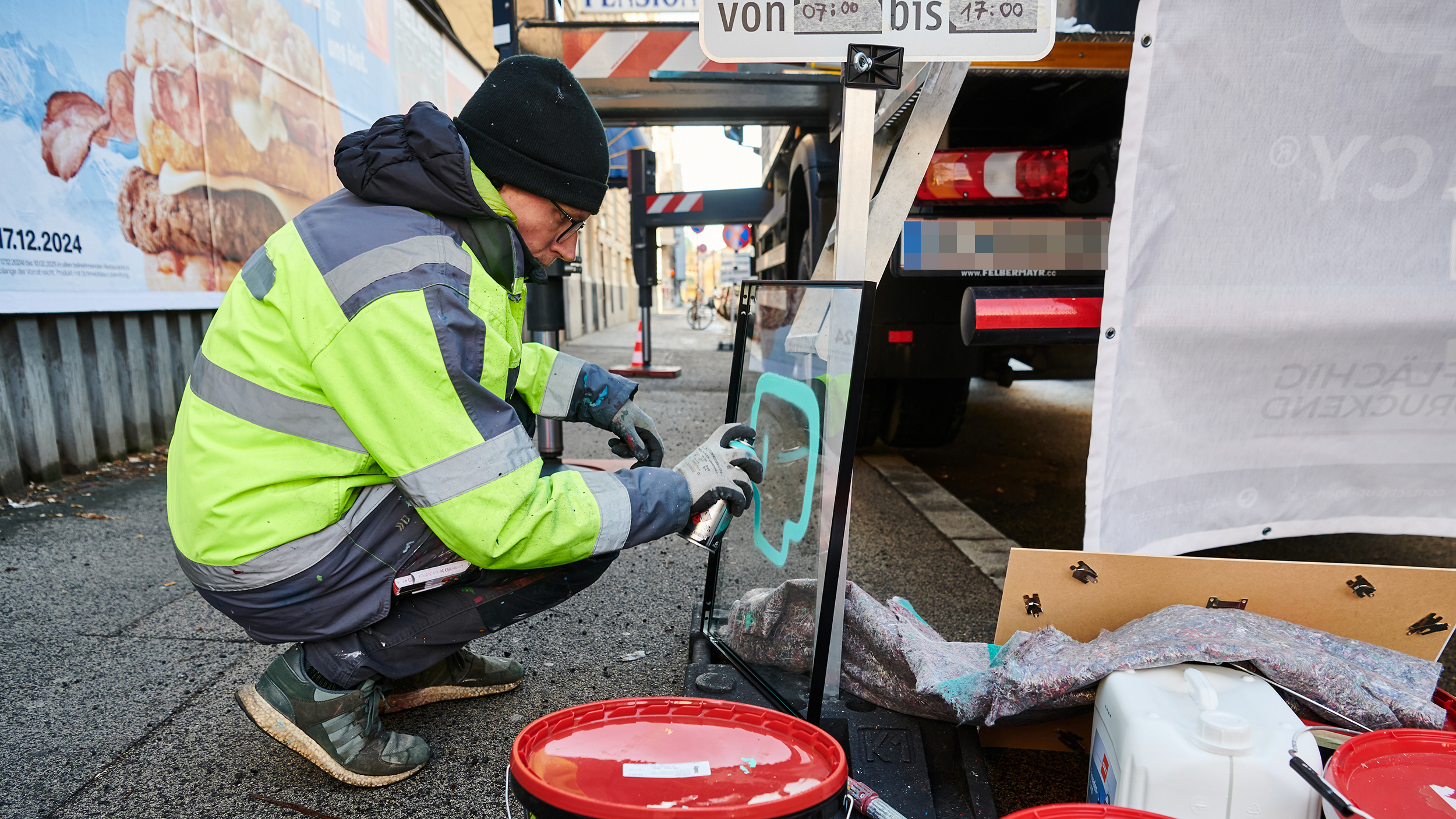 Graffiti-Künster sizetwo sprüht auf eine Glasplatte etwas auf. Er hockerlt dabei neben einem LKW am Gehsteig.