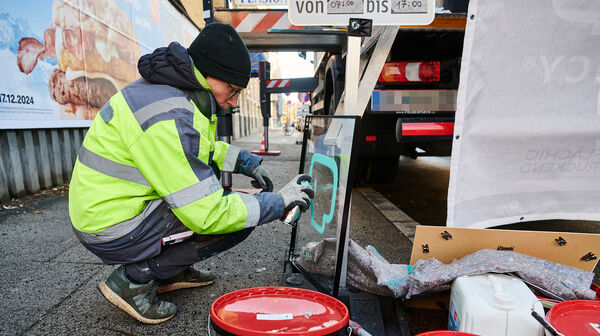 Graffiti-Künster sizetwo sprüht auf eine Glasplatte etwas auf. Er hockerlt dabei neben einem LKW am Gehsteig.