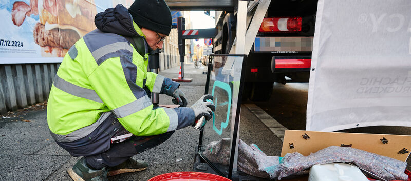 Graffiti-Künster sizetwo sprüht auf eine Glasplatte etwas auf. Er hockerlt dabei neben einem LKW am Gehsteig.