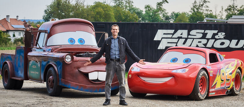 Martin Hahn steht vor den Filmautos aus Cars, Lightning McQueen (links) und Hook (rechts)
