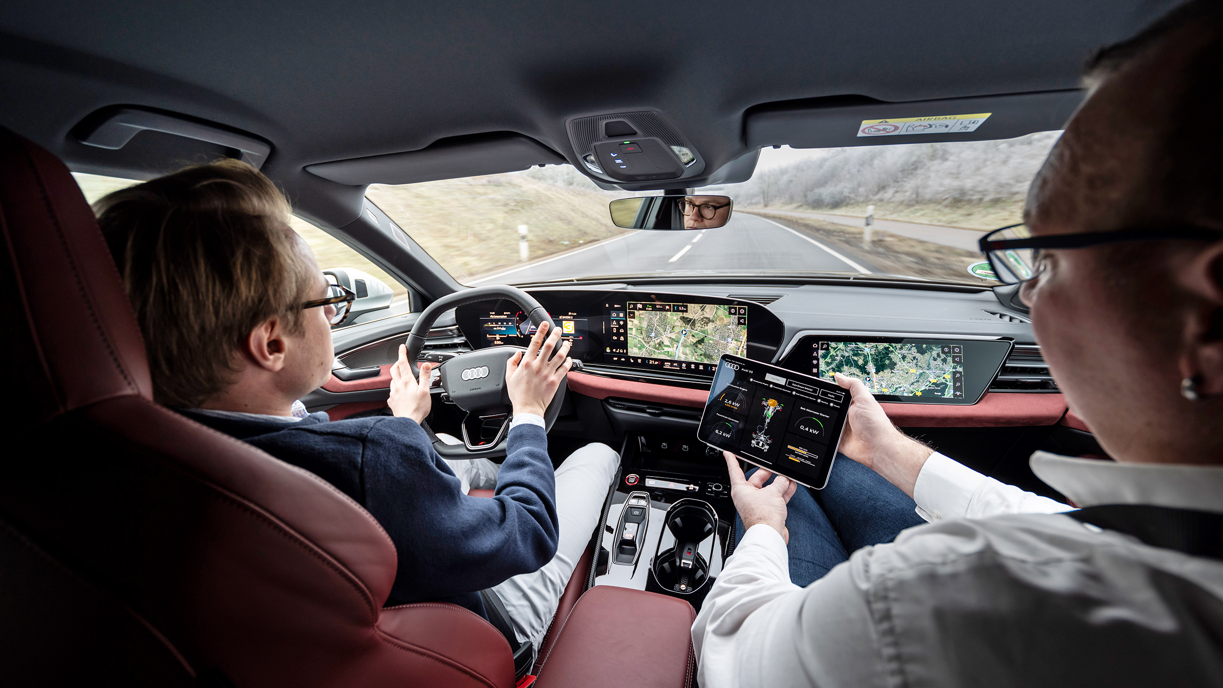 Zwei Personen sitzen in einem Auto, die linke Person fährt, die rechte Person hält ein Tablet mit verschiedenen Fahrdaten.