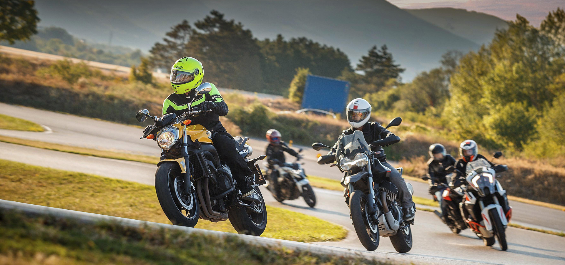 Vier Personen auf Motorrädern fahren hinter einem Instruktor auf einem gelben Motorrad. Im Hintergrund sind grüne Hügel, asphaltierte Straßen und Bäume.