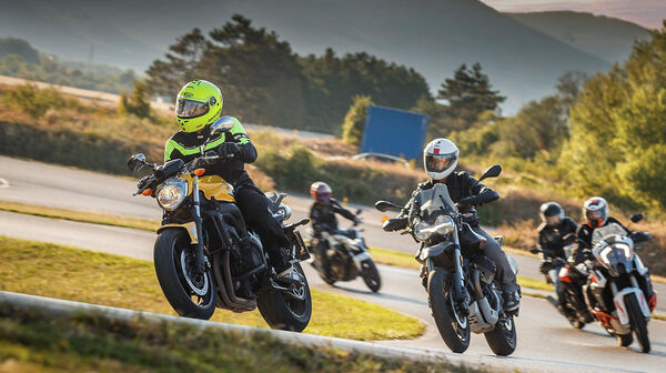 Vier Personen auf Motorrädern fahren hinter einem Instruktor auf einem gelben Motorrad. Im Hintergrund sind grüne Hügel, asphaltierte Straßen und Bäume.
