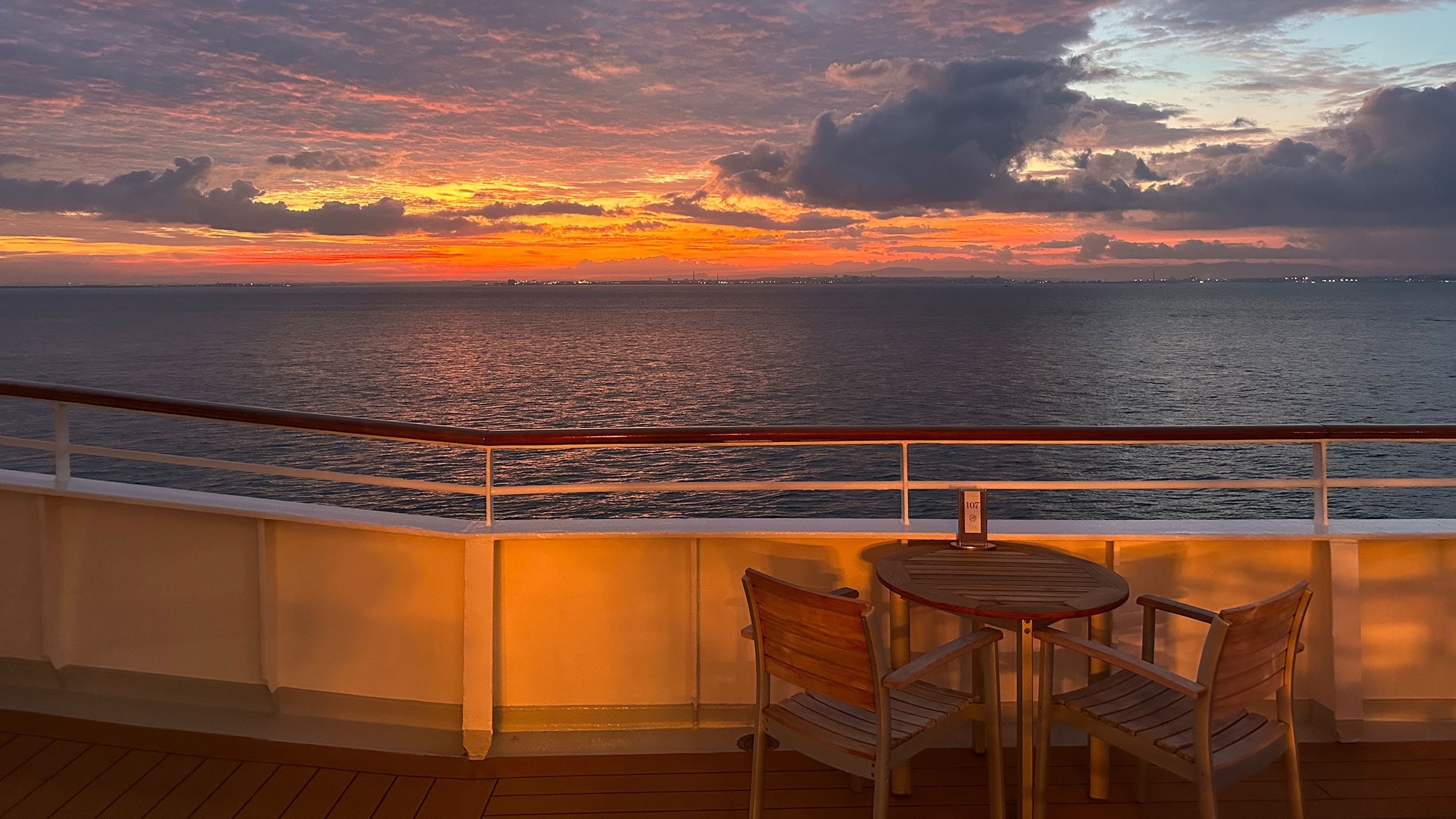 Zu sehen ist ein Tisch und zwei Stühle auf Deck an der Reling der MS Europa bei Sonnenuntgergang mit Blick auf das Meer.
