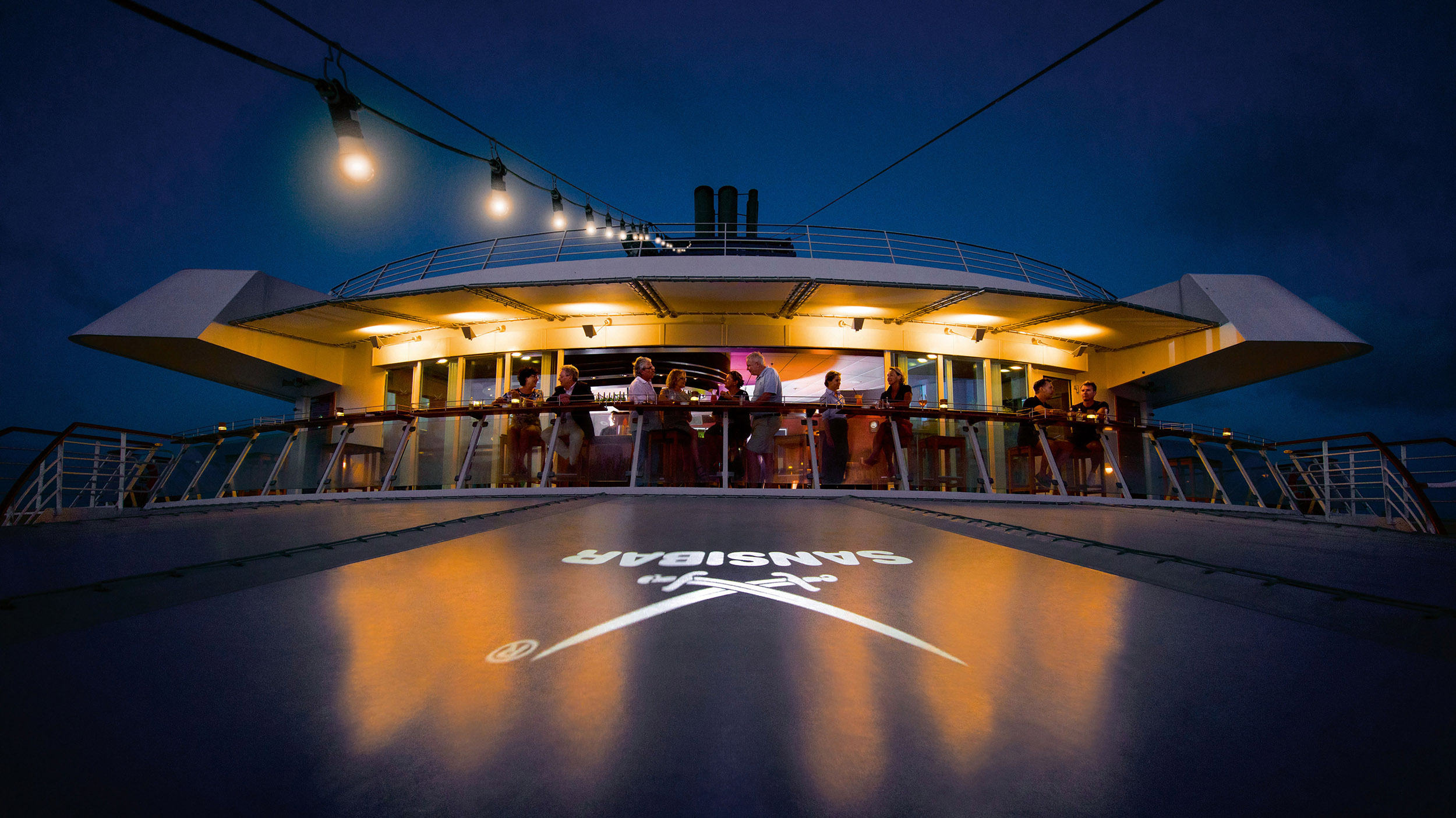 Einige Menschen stehen in der Nacht an Deck der Bar "Sansibar" auf der MS Europa und genießen den Abend.