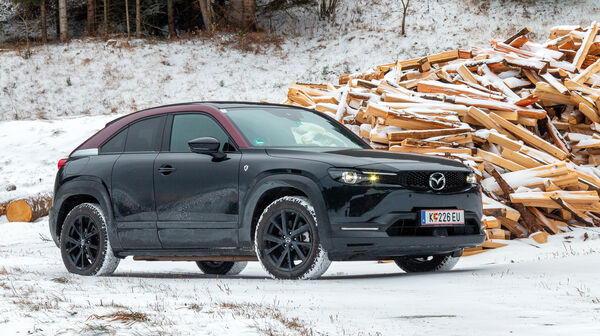 Ein schwarz-roter Mazda MX-30 steht in einer verschneiten Winterlandschaft vor einem Haufen Holz.