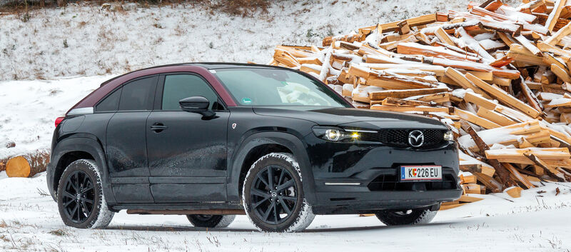 Ein schwarz-roter Mazda MX-30 steht in einer verschneiten Winterlandschaft vor einem Haufen Holz.