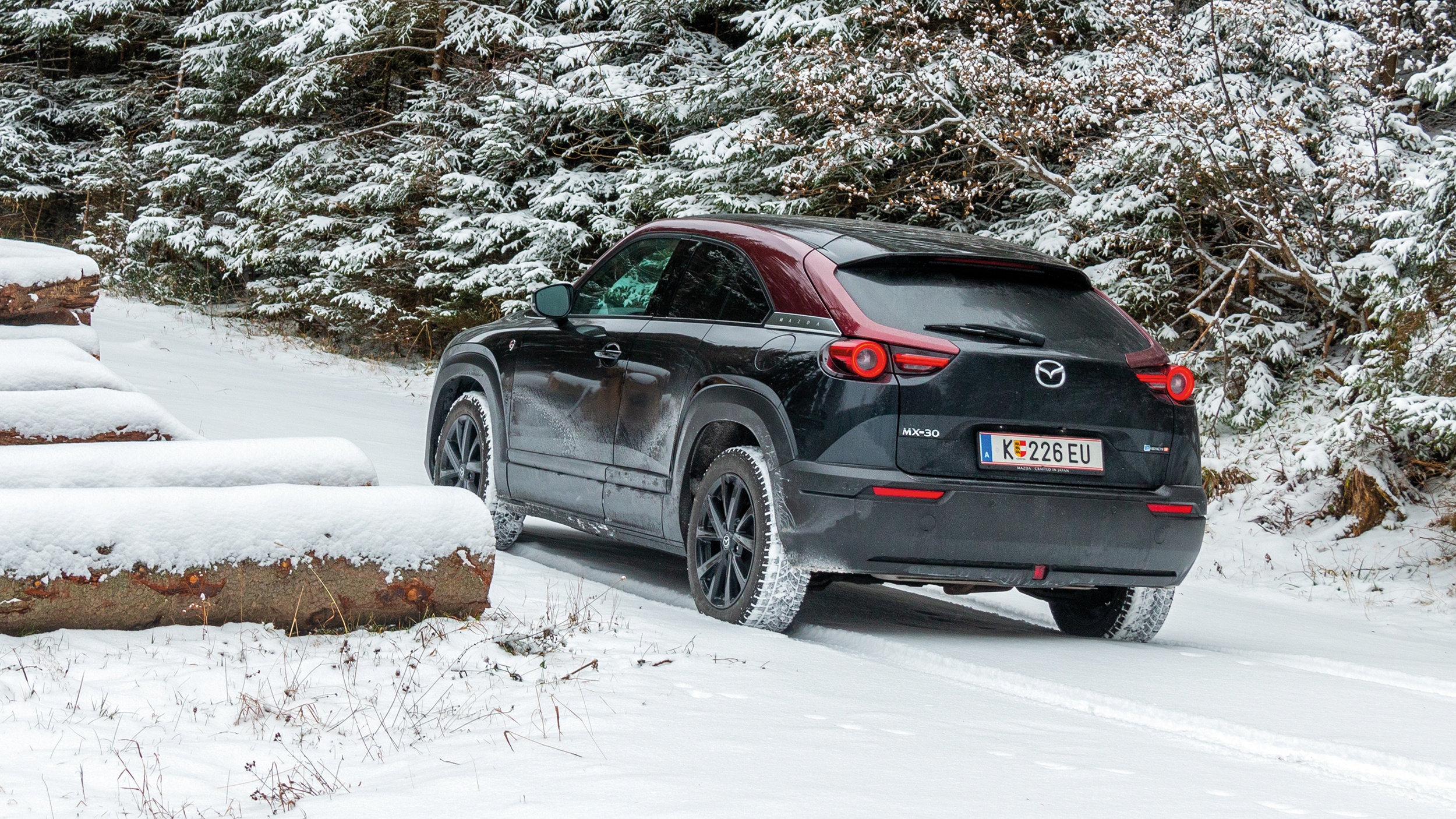 Ein schwarz-roter Mazda MX-30 steht im verschneiten Wald zwischen Bäumen und abgefällten Baumstämmen.