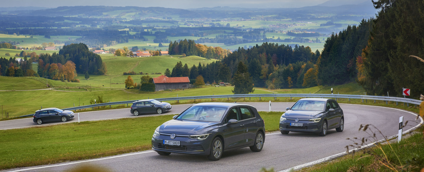 In einer ländlichen Umgebung fahren 4 Autos eine kurvige Straße bergauf. Im Hintergrund Wälder, Häuser und Wiesen.
