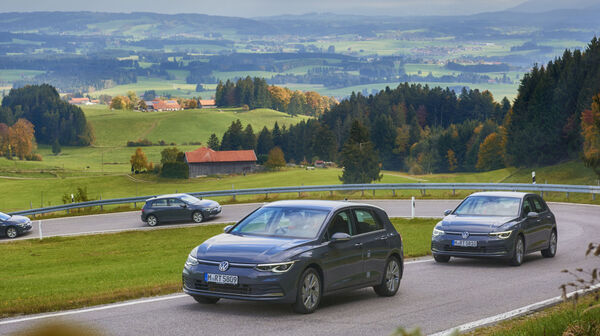 In einer ländlichen Umgebung fahren 4 Autos eine kurvige Straße bergauf. Im Hintergrund Wälder, Häuser und Wiesen.