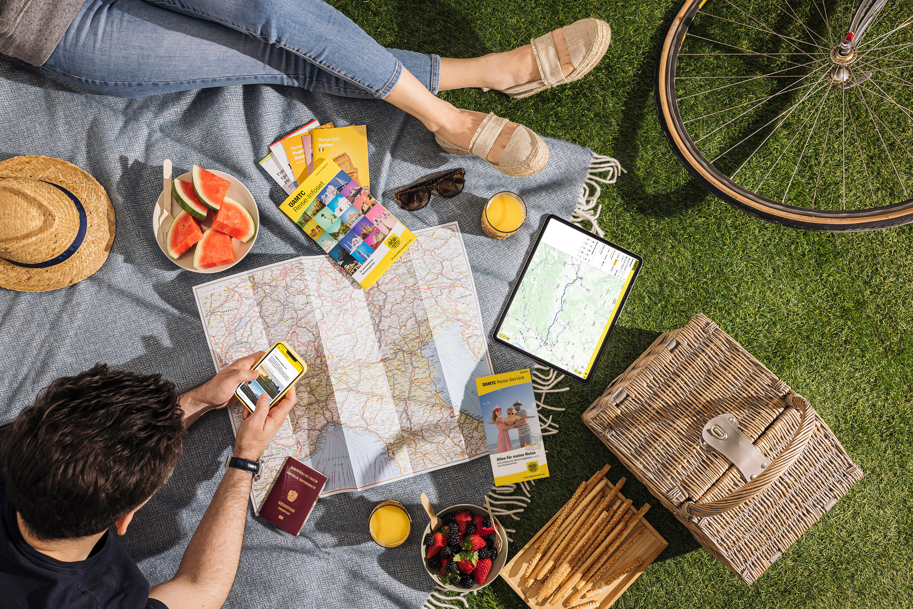Zwei Personen sitzen auf einer Picknickdecke im Gras. Auf der Decke liegen zudem Landkarten, ein Tablet, Reiseführer, Obst, ein Hut. Im Eck sieht man den Reifen eines Fahrrads.
