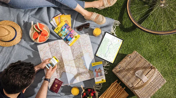 Zwei Personen sitzen auf einer Picknickdecke im Gras. Auf der Decke liegen zudem Landkarten, ein Tablet, Reiseführer, Obst, ein Hut. Im Eck sieht man den Reifen eines Fahrrads.