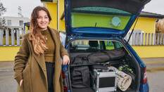 Sängerin Anna Pflamitzer posiert vor ihrem blauen Auto, das vor einem gelben Haus geparkt ist. Der Kofferraum ist geöffnet und zeigt ihr Arbeitsmaterial.