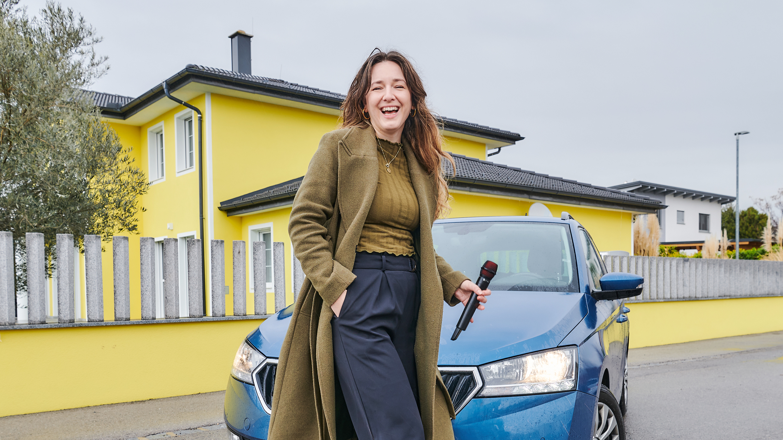 Sängerin Anna Pflamitzer steht lachend vor ihrem blauen Auto, das vor einem gelben Haus geparkt ist. In der Hand hält sie ein Mikrophon. 