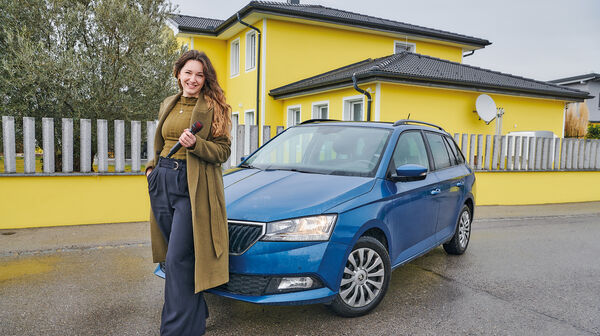 Sängerin Anna Pflamitzer posiert vor ihrem blauen Auto, das vor einem gelben Haus geparkt ist. In der Hand hält sie ein Mikrophon.