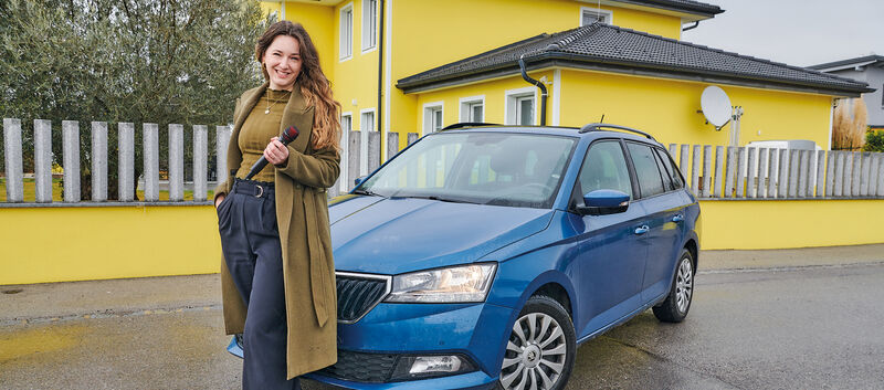 Sängerin Anna Pflamitzer posiert vor ihrem blauen Auto, das vor einem gelben Haus geparkt ist. In der Hand hält sie ein Mikrophon.