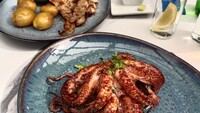 Es ist ein Foto von einem Tisch in einem Restaurant auf dem zwei Teller stehen. Auf einem Teller ist ein Fleischgericht mit Kartoffeln zu sehen, auf dem blauen Teller im Vordergrund des Bildes ist ein gebratener Tintenfisch.