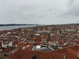 Das Foto zeigt Lissabon aus der Vogelperspektive - es sind viele rote Dächer von unterschiedlichen Gebäuden der Stadt zu sehen. Im linken Bildrand kann man auch das Meer erkennen.