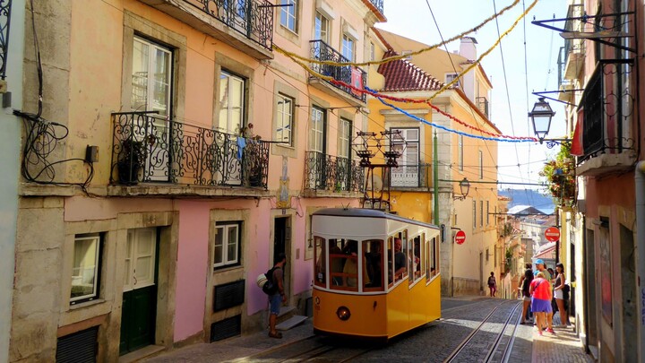 Auf dem Foto ist eine enge Straße in Lissabon zu sehen, links und rechts ist jeweils eine Häuserfront und in der Mitte fährt die berühmte Standseilbahn.