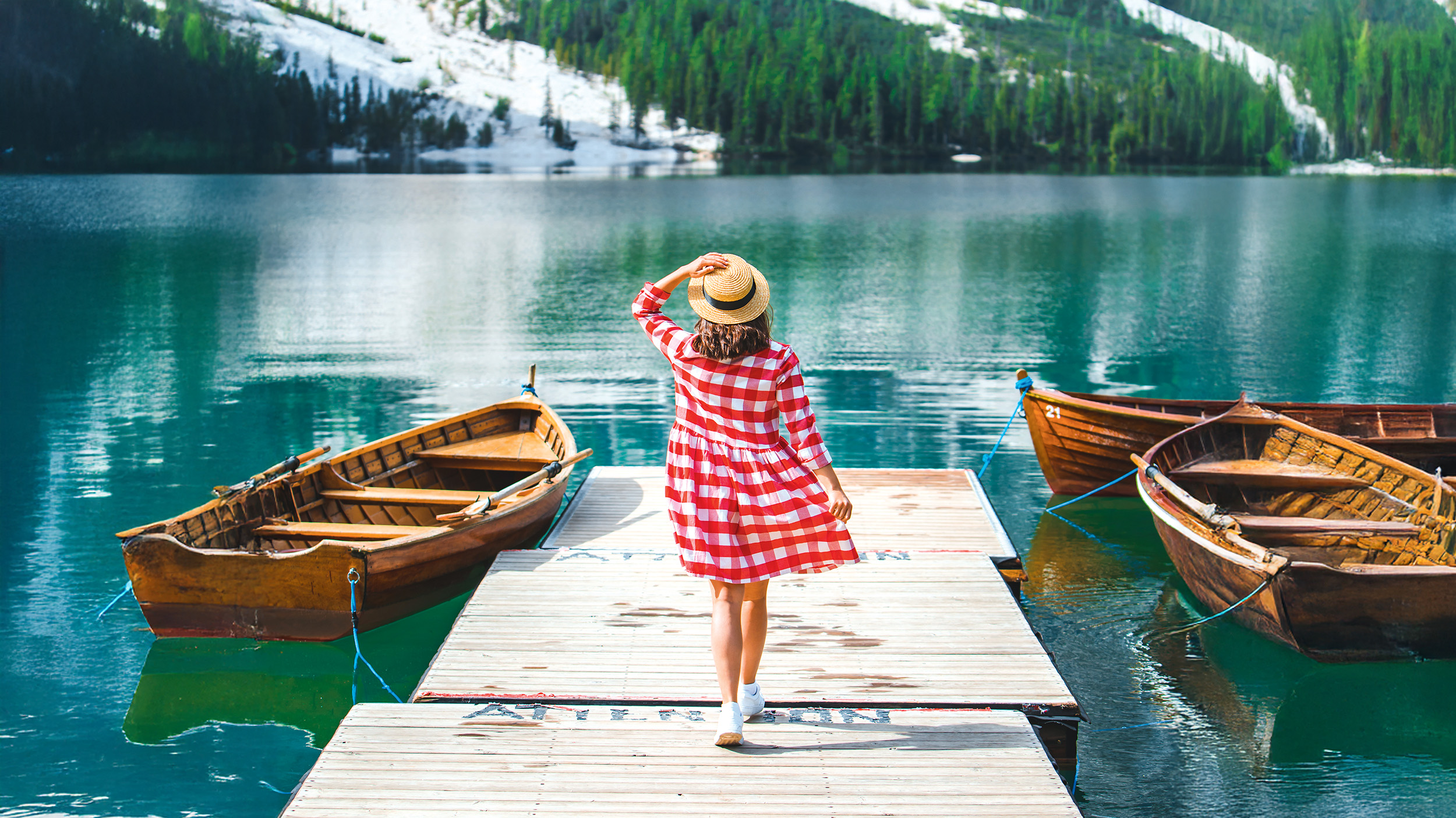 Eine Frau mit Hut in einem rot-weiß karierten Kleid geht über einen Steg, an dem Ruderboote liegen.