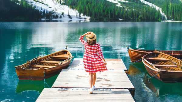 Eine Frau mit Hut in einem rot-weiß karierten Kleid geht über einen Steg, an dem Ruderboote liegen.