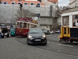 Auf dem Bild ist ein Straße auf der ein schwarzes Auto zwischen zwei Straßenbahnen fährt zu sehen. die hintere Straßenbahn ist rot, die Straßenbahn auf der rechten Seite des Bildes ist gelb. Am Straßenrand, im Hintergrund des Bildes, sind auch mehrere Tuk-Tuks zu sehen.