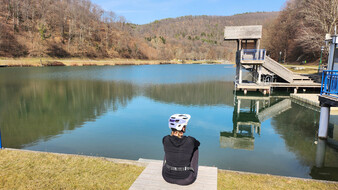 Redakteurin Verena Schauer am Badesee in Rechnitz. Sie sitzt am Steg, man sieht sie von hinten. Sie trägt einen Fahrradhelm und ist schwarz gekleidet.