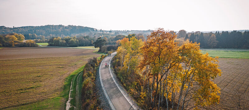 Bahntrassen-Radweg aus der Vogelperspektive. Es ist eine herbstliche Wettersituation, man sieht klein ein paar Fahrradfahrer:innen.