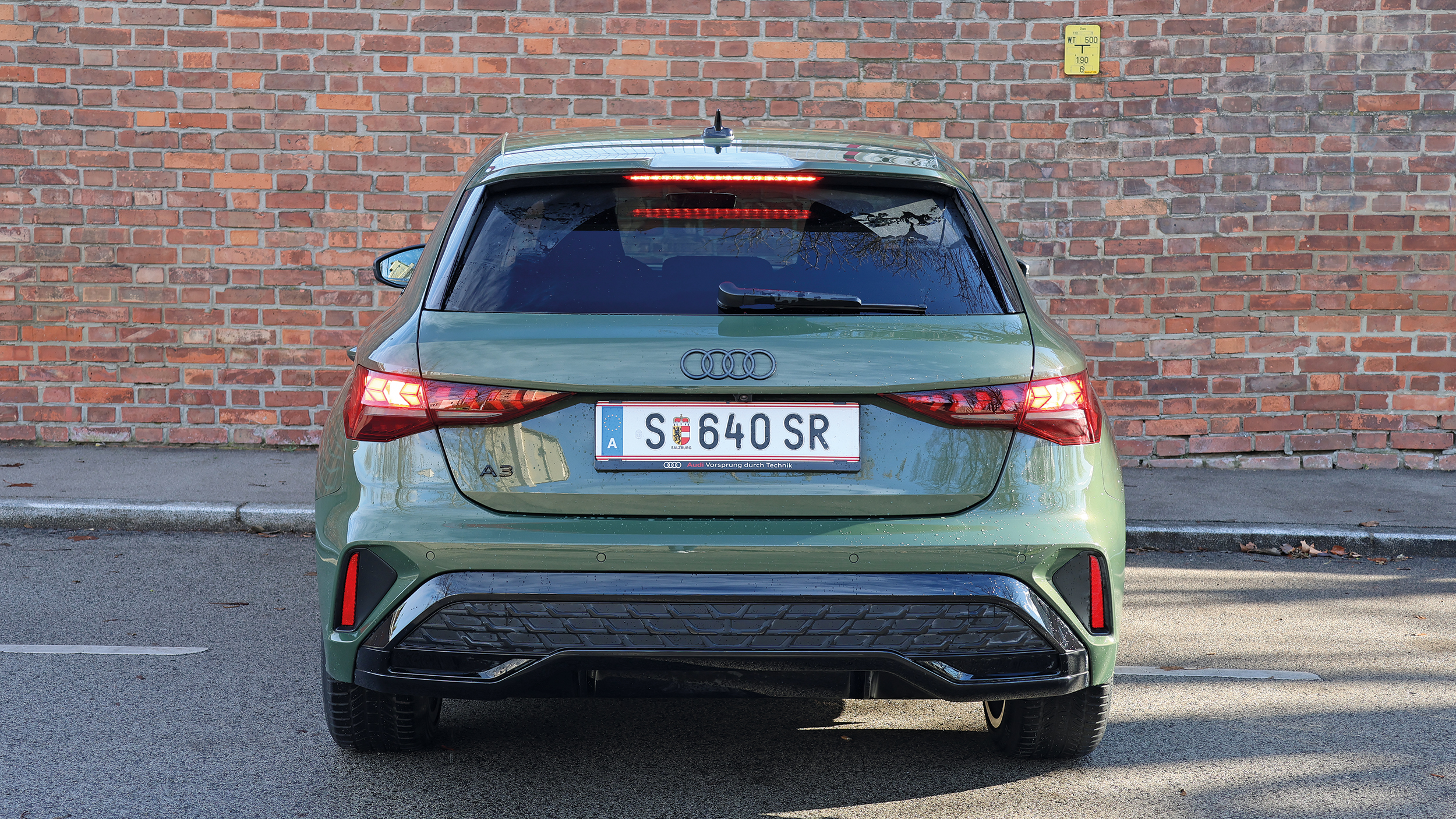 Zu sehen ist die Heckansicht eines grünen Audi A3 Sportback vor einer Backsteinmauer.