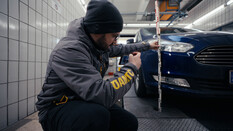 Auf dem Bild ist ein Schadensbeguachter bei der Arbeit zu sehen. Der Mann trägt eine dunkelgraue Jacke mit ÖAMTC Logo und hat eine graue Mütze auf dem Kopf. Er kniet neben einem blauen Auto und hält einen Zollstock und misst die Höhe vom Boden bis zu den Scheinwerfern auf der Vorderseite des Fahrzeugs.