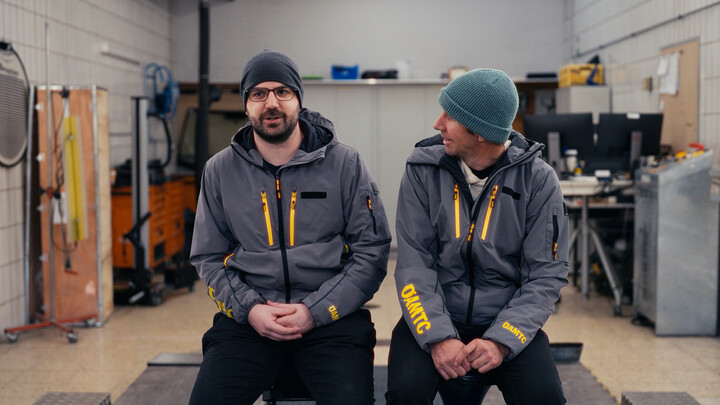 Auf dem Bild sind zwei Männer zu sehen, die nebeneinander in einer Werkstatt sitzen. Beide tragen eine dunkelgraue Jacke mit ÖAMTC-Logo. Der linke Mann schaut in die Kamera und trägt eine dunkelgraue Mütze, der rechte Mann schaut den anderen an und trägt eine blaue Mütze.