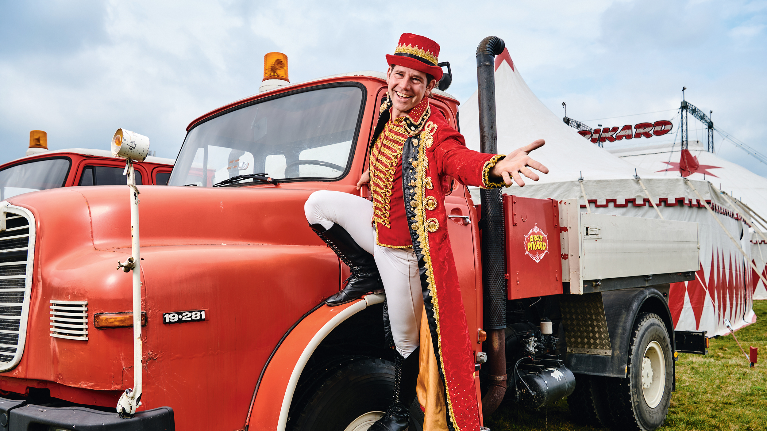 Ein Mann in einem Rot-weißen Kostüm eines Zirkusdirektors steht am Kotflügel eines alten, roten MAN-Zugfahrzeugs. In Hintergrund das Zirkuszelt.