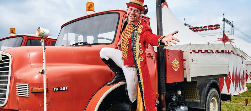 Ein Mann in einem Rot-weißen Kostüm eines Zirkusdirektors steht am Kotflügel eines alten, roten MAN-Zugfahrzeugs. In Hintergrund das Zirkuszelt.