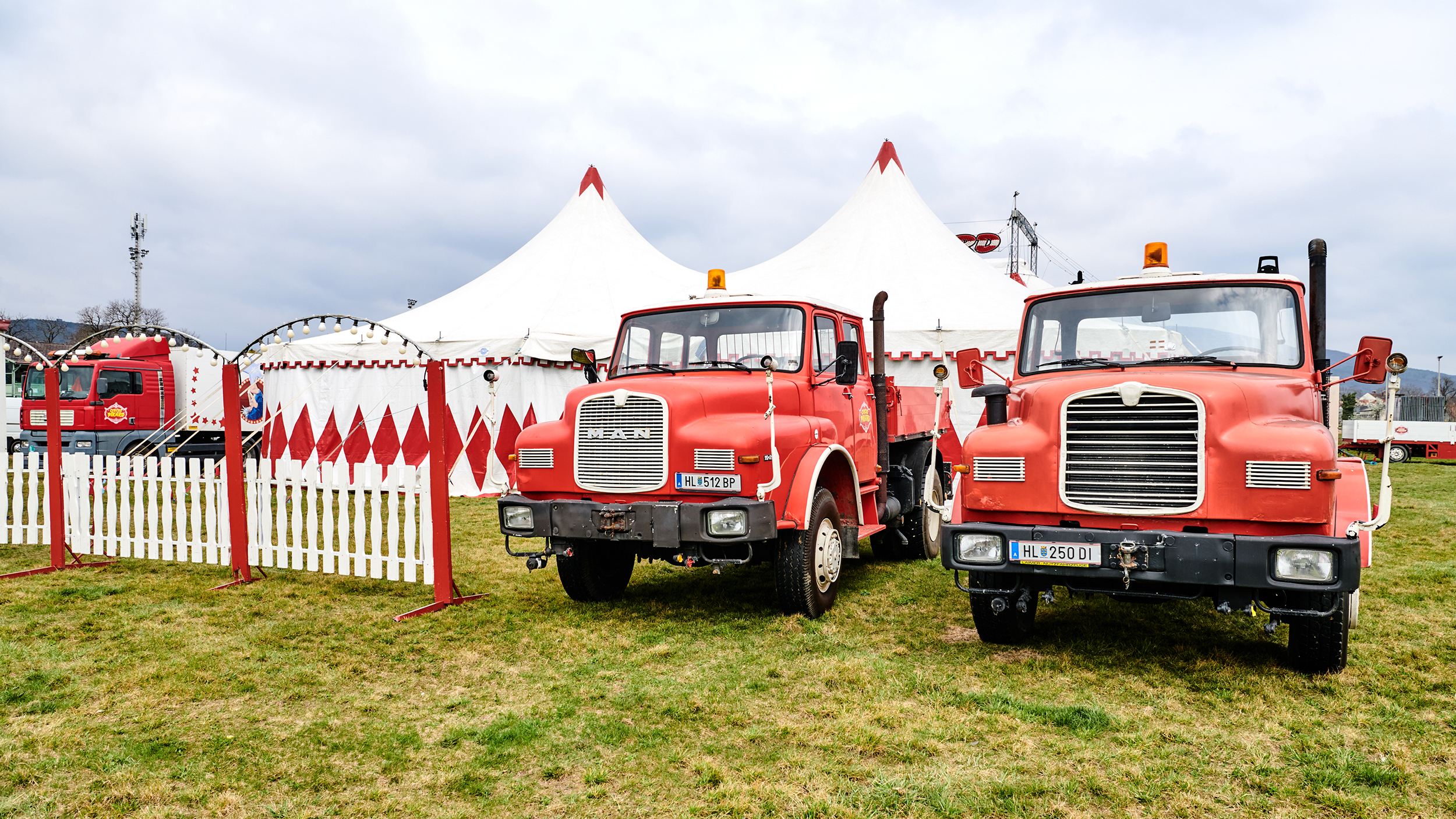 Zwie alte rote Zugfahrzeuge stehen neben einem weißen Zaun auf einer Wiese, im Hintergrund ein Zirkuszelt.