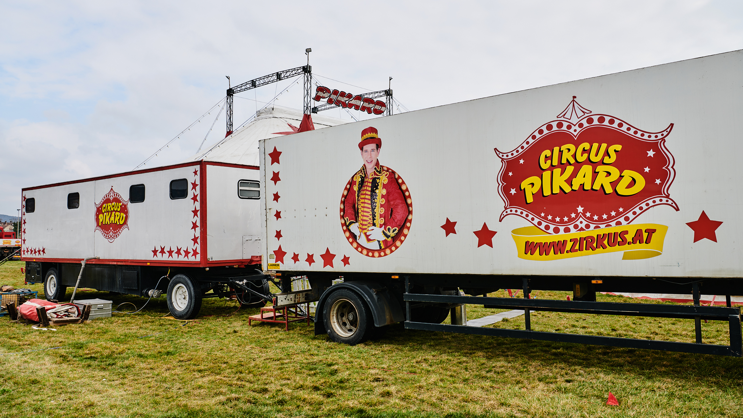 Zwei weiße LKW-Anhänger mit Bild des Zirkusdirektors und Logo vom Cirkus Pikard stehen auf einer Wiese. 