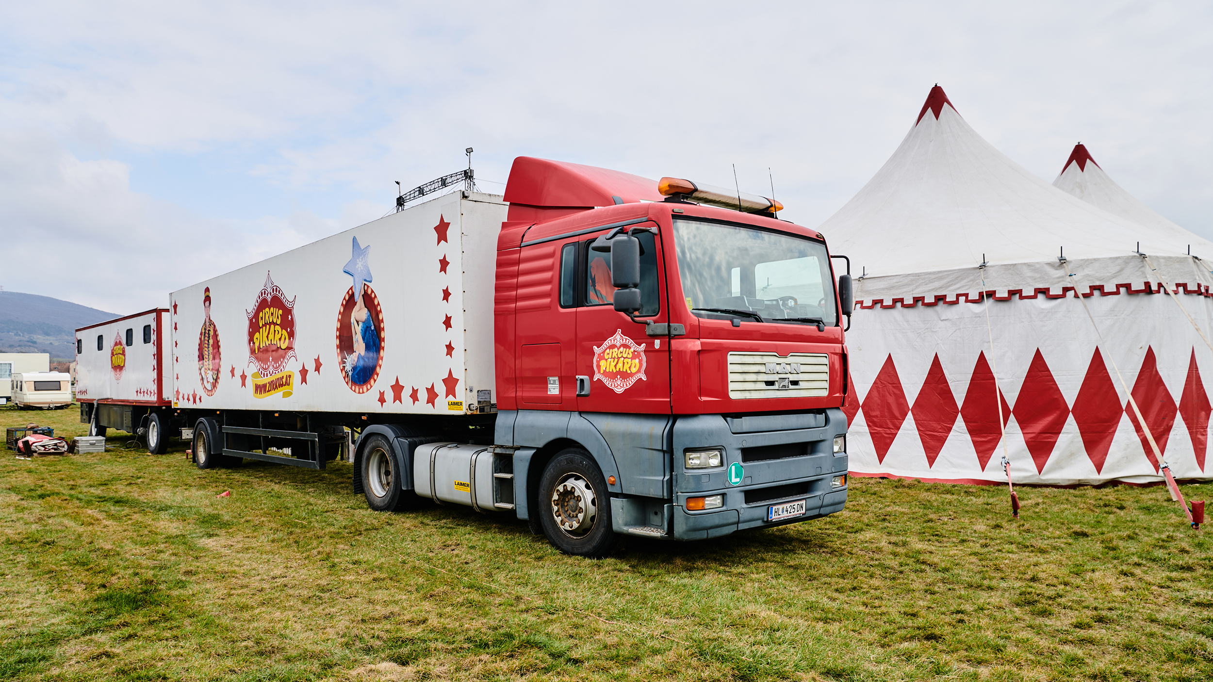 Ein rotes LKW-Zugfahrzeug mit 2 Anhängern des Circus Pikard steht vor dem Zirkuszelt.
