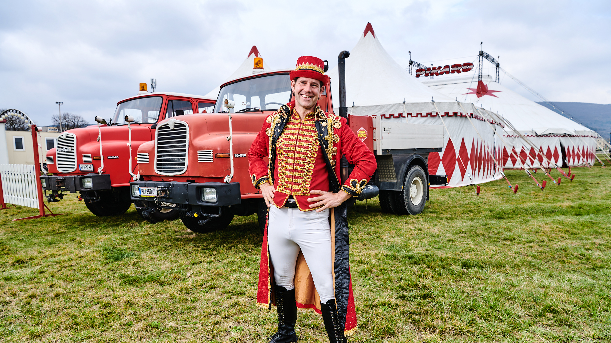 Zirkusdirektor Alexander Schneller steht im Kostüm vor seinen Zugfahrzeugen und dem Zirkuszelt.