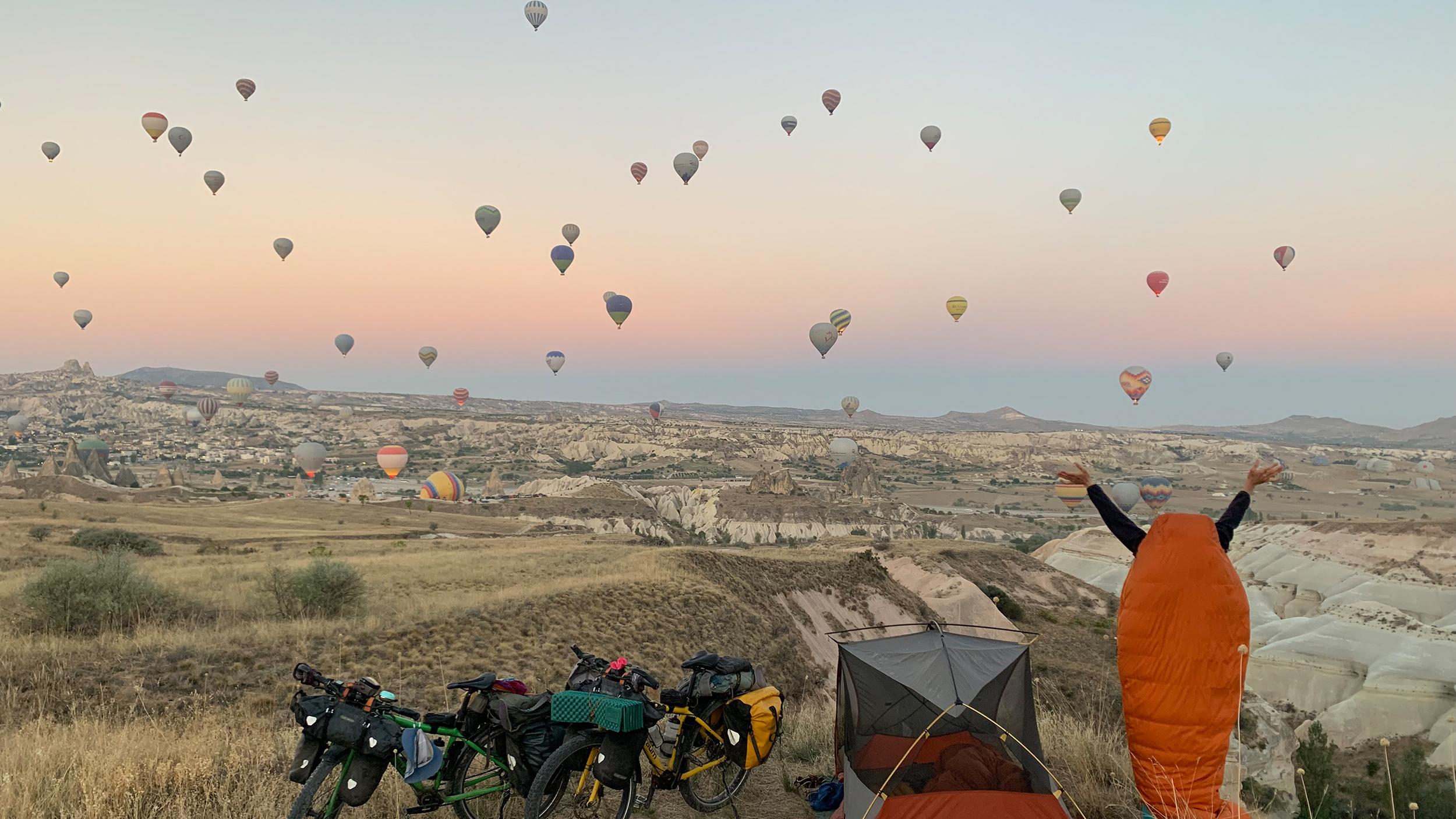 Eine hügelige grüne Landschaft mit unzähligen Heißluftballons in Kappadokien im Hintergrund. Im Vordergrund zwei Räder, ein Zelt und eine Person steht im Schlafsack und blickt auf die Ballons.