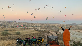 Eine hügelige grüne Landschaft mit unzähligen Heißluftballons in Kappadokien im Hintergrund. Im Vordergrund zwei Räder, ein Zelt und eine Person steht im Schlafsack und blickt auf die Ballons.
