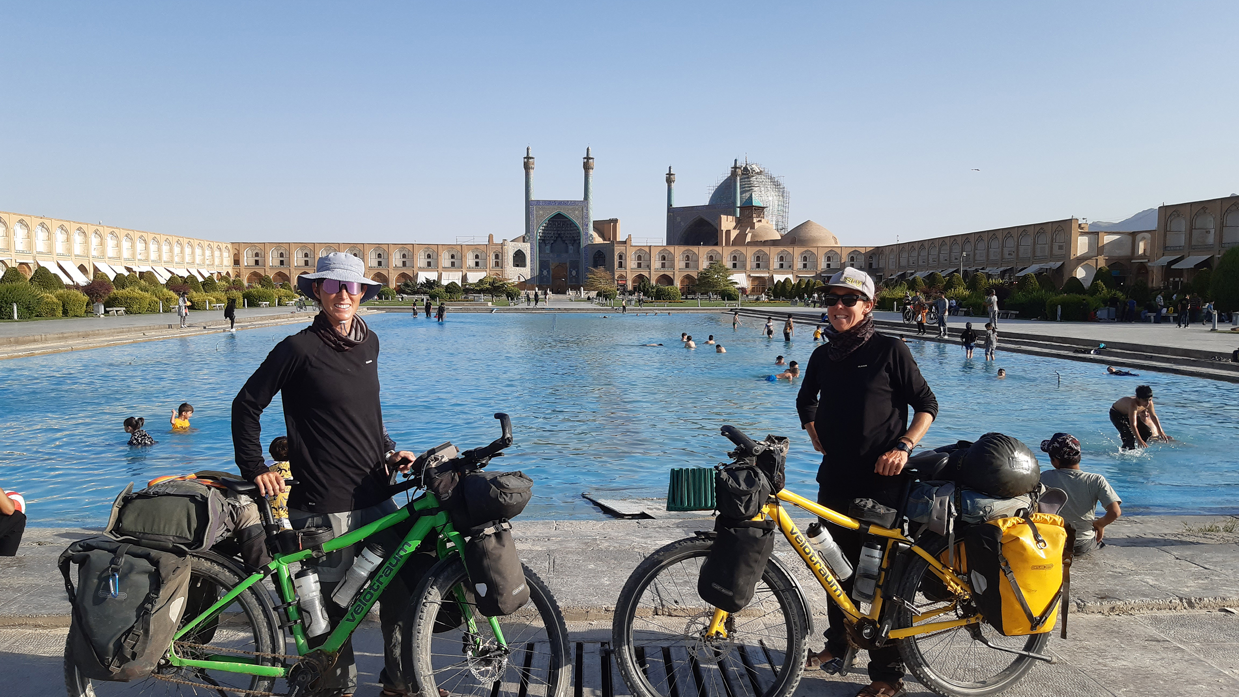 Zwei Frauen mit Hut und Sonnenbrille stehen mit ihrem bepackten Rädern vor dem Wasserbassin der Moschee in Isfahan, die im Hintergrund zu sehen ist.
